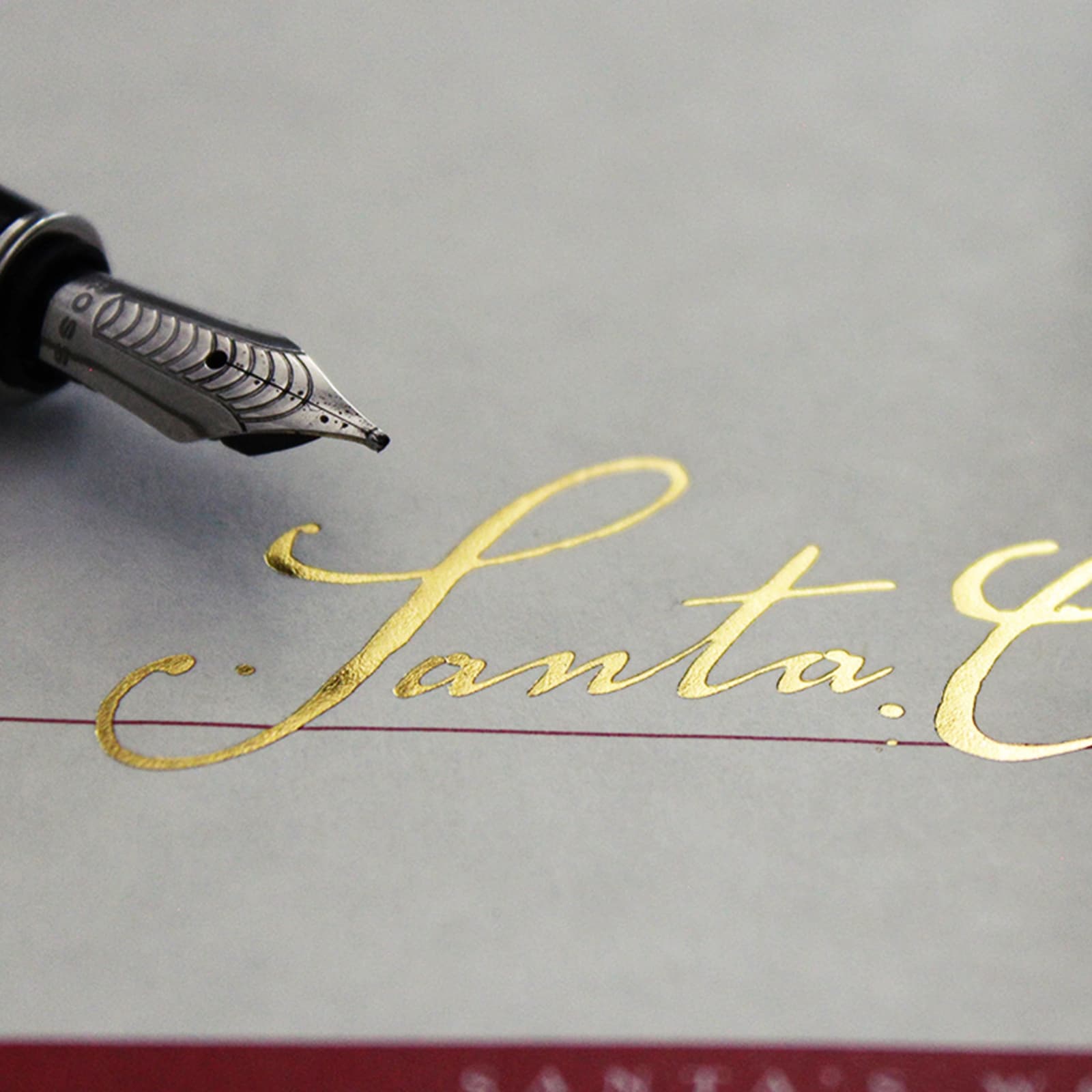Close-up macro of the gold-foil 'Santa' signature on the Red Letter, with a fountain pen above and the red Santa's Red Letter wordmark visible at the bottom.